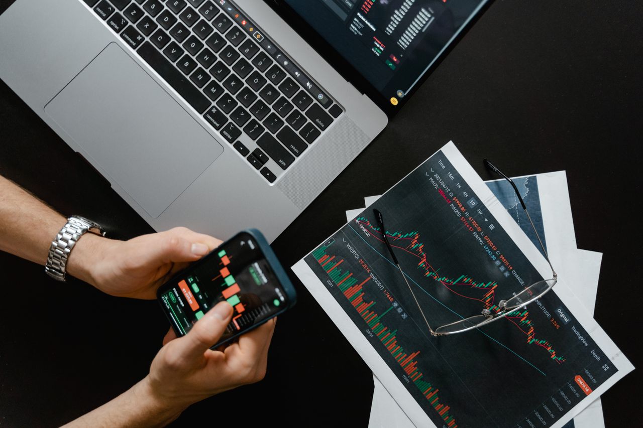 services-02 Des mains utilisant un smartphone à côté d’un ordinateur portable avec des graphiques boursiers, illustrant le trading numérique.